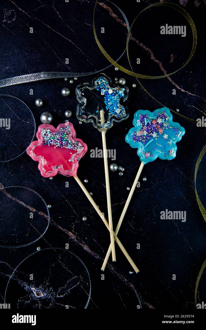 Lollipops on a wooden stick in the form of a rattle in the form of transparent, pink and blue snowflakes lie on a dark marble background surrounded by gold and silver ribbons and silver balls, vertical photography Stock Photo