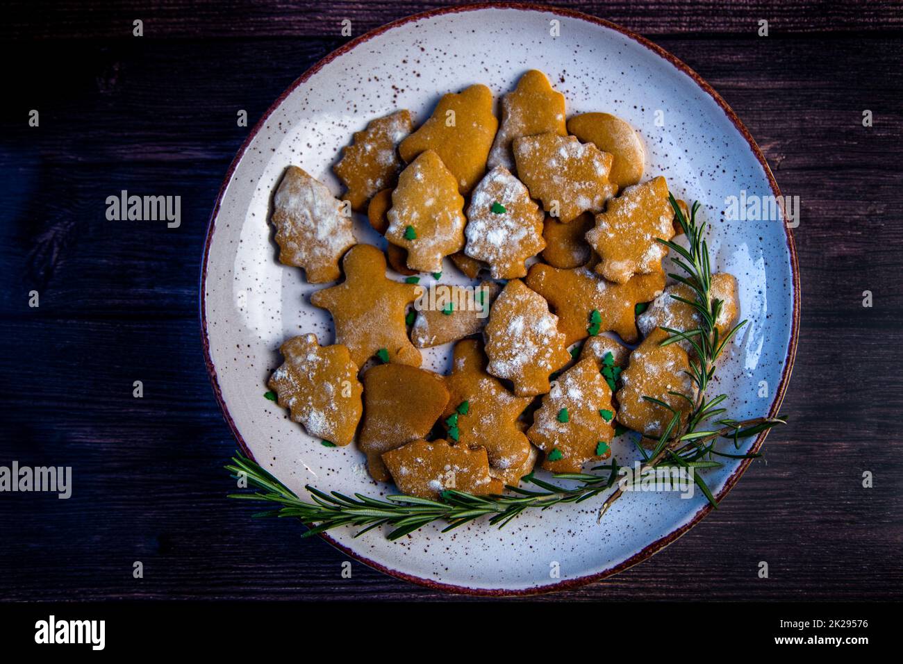 Cookies are on a plate of different shapes, in the foreground there is a cookie in the form of a Christmas tree, decorated with powdered sugar like snow and small green Christmas trees against the background of the rest of the cookies, a branch of rosemar Stock Photo