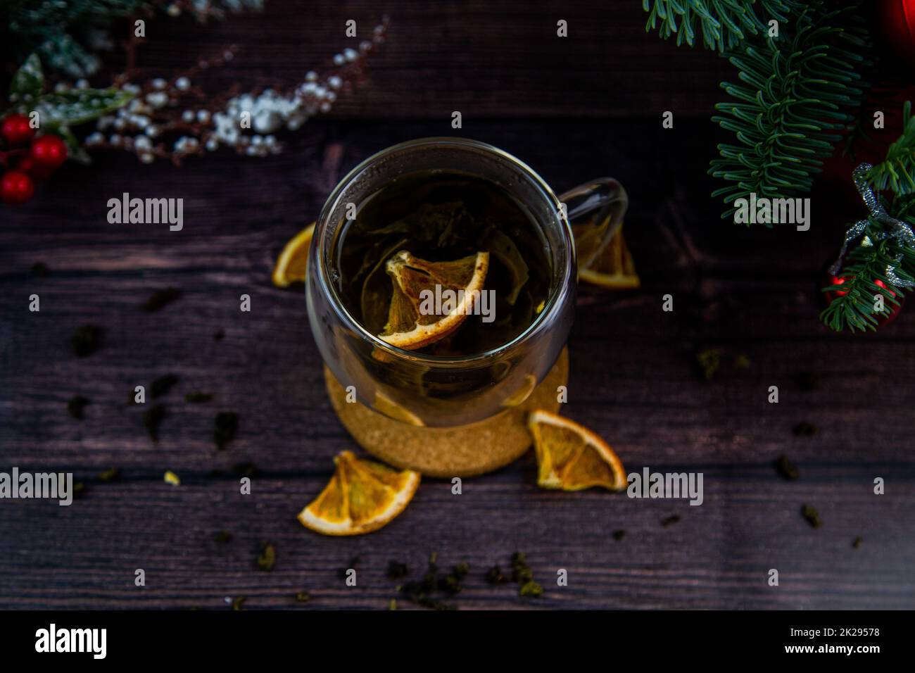 A transparent cup of tea stands on a dark table, on a wooden stand, dried orange slices float in the cup, citrus slices lie next to it, tea is scattered, a photo in a dark key Stock Photo