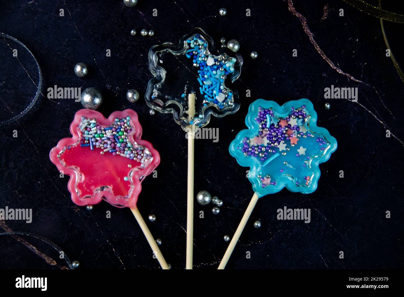 Lollipops on a wooden stick in the form of a rattle in the form of transparent, pink and blue snowflakes lie on a dark marble background surrounded by gold and silver ribbons and silver balls Stock Photo