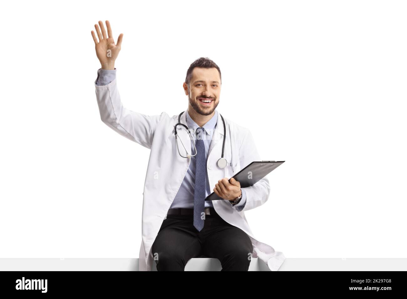 Young male doctor sitting on a blank panel and waving isolated on white background Stock Photo