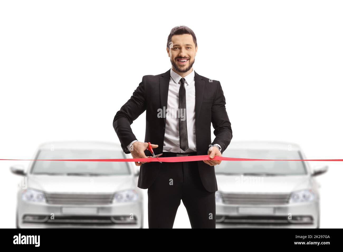 Manager in a suit cutting a red ribbon tape in a car showroom and smiling at the camera isolated on white background Stock Photo