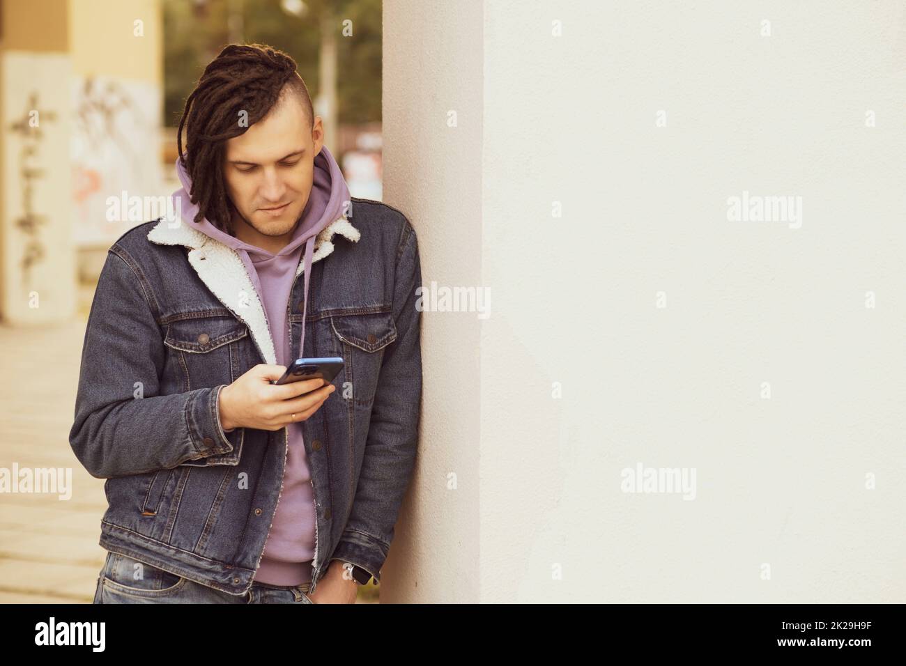 Portrait of young handsome man with dreadlocks browsing mobile phone, standing, leaning against wall on street Stock Photo