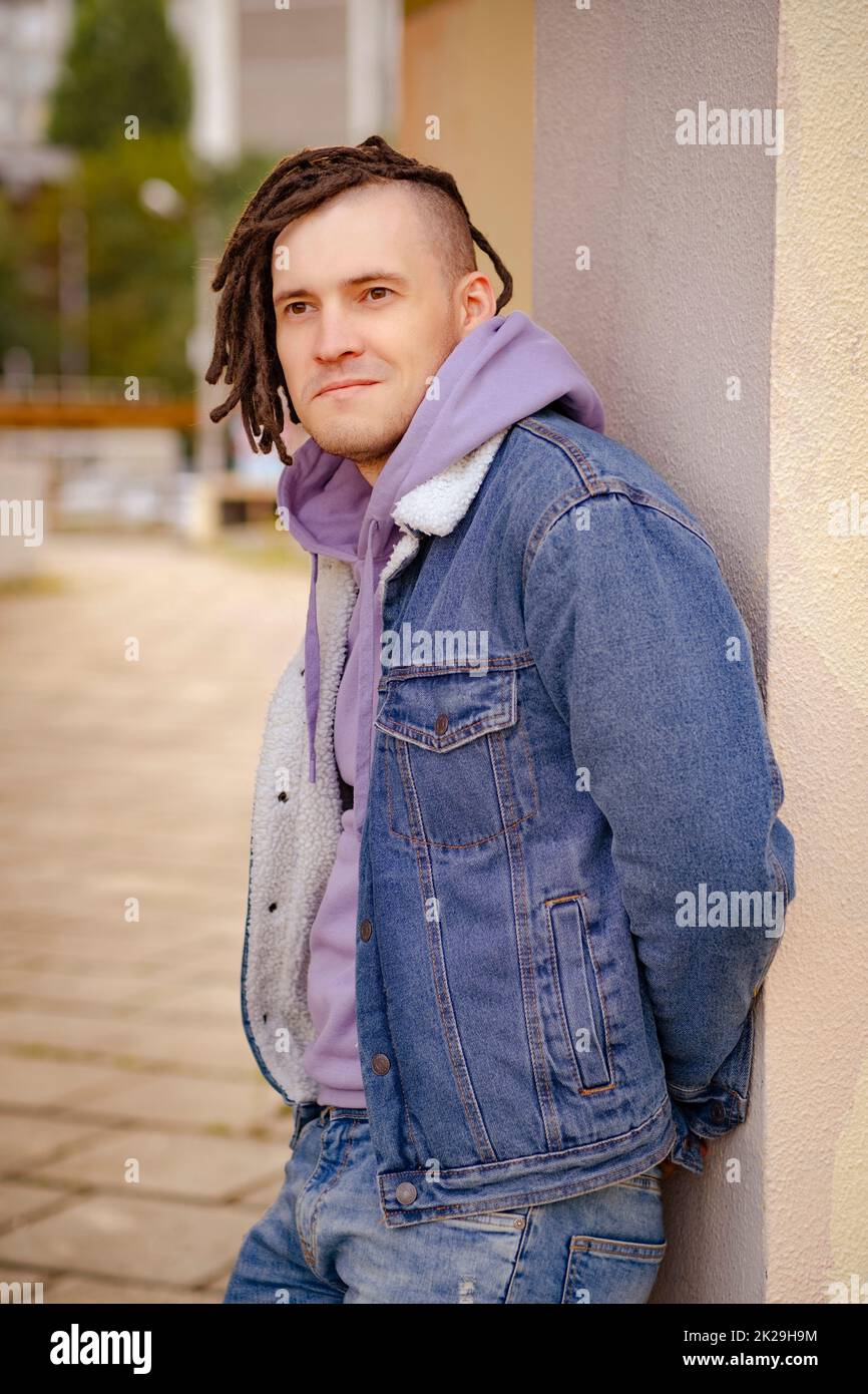 Portrait of young handsome man with dreadlocks posing, standing, leaning against wall on street Stock Photo