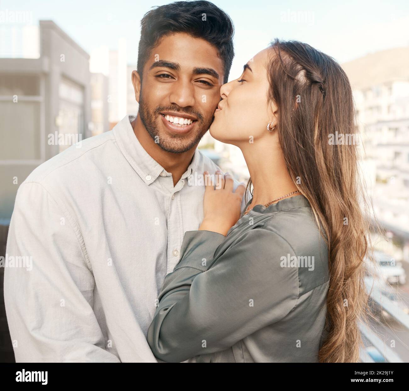 Happy couple, love and kiss on cheek from girlfriend bonding with boyfriend standing on a city building balcony on a holiday. Portrait of indian man Stock Photo