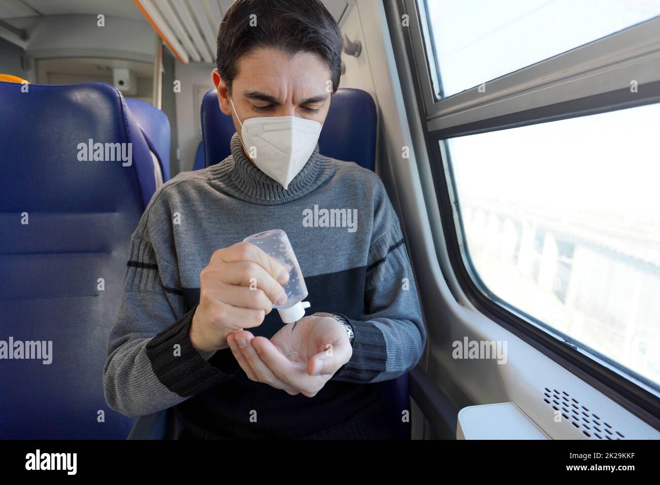 Travel safely on public transport. Young man with protective face mask disinfects hands from alcohol gel dispenser. Passenger with medical mask sanitizing hands inside train carriage. Stock Photo