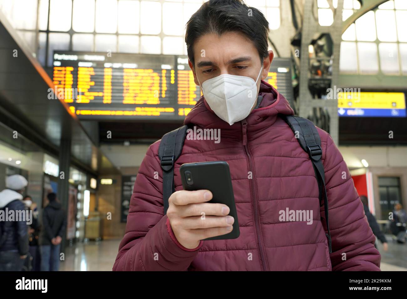 Traveler man wearing KN95 FFP2 protective face mask at the airport. Young caucasian man with behind timetables board of departures arrivals checking online ticket of his flight. Stock Photo