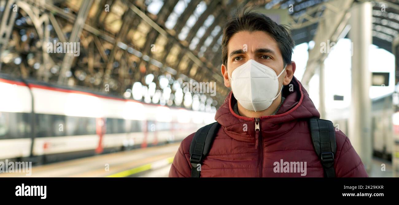 Banner panorama of young traveler man wearing protective face mask at train station with copy space. Public transport and health concept. Stock Photo