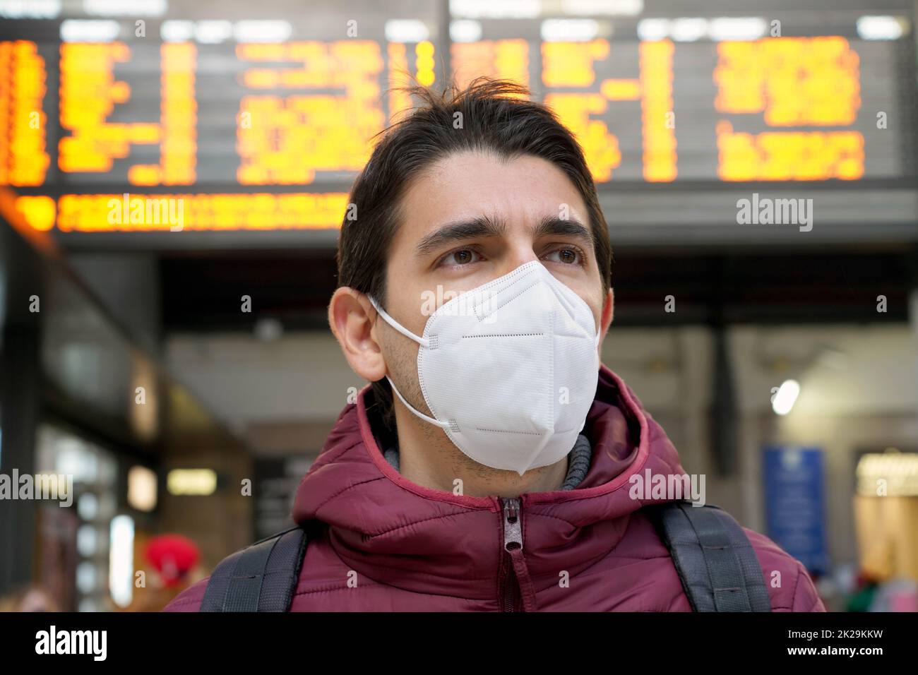 Close-up traveler man wearing KN95 FFP2 face mask at the airport. Young caucasian man with behind timetables board of departures arrivals waiting worried for information on his flight. Stock Photo