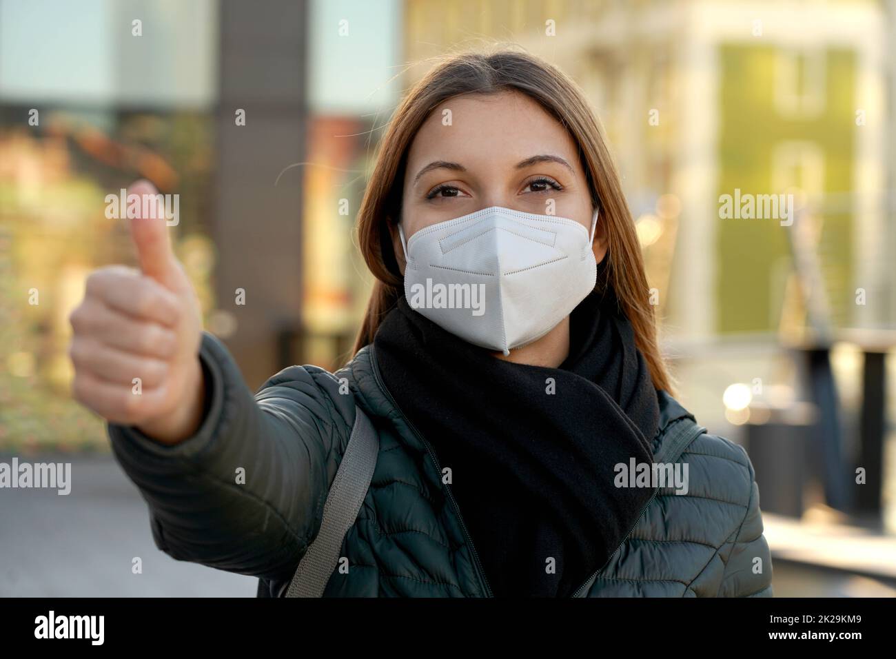 Happy young woman wearing medical face mask showing thumbs up in the evening outside. Looking camera. Stock Photo
