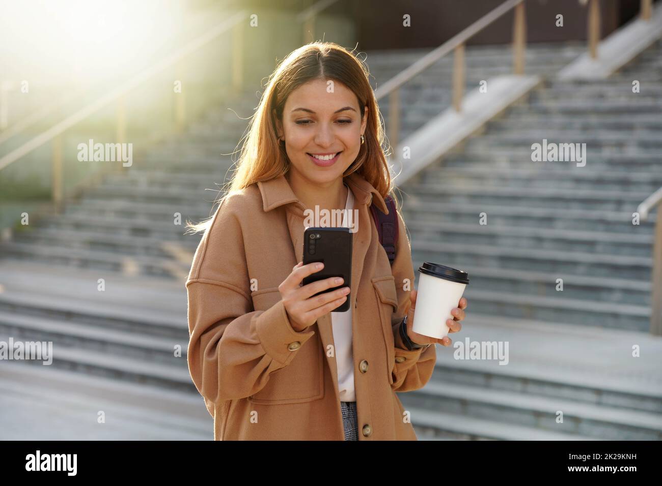 Enchanting blonde young woman waiting for phone message while carrying coffee cup on the street. Stylish girl in spring clothing holding smartphone and take-away cappuccino. Stock Photo
