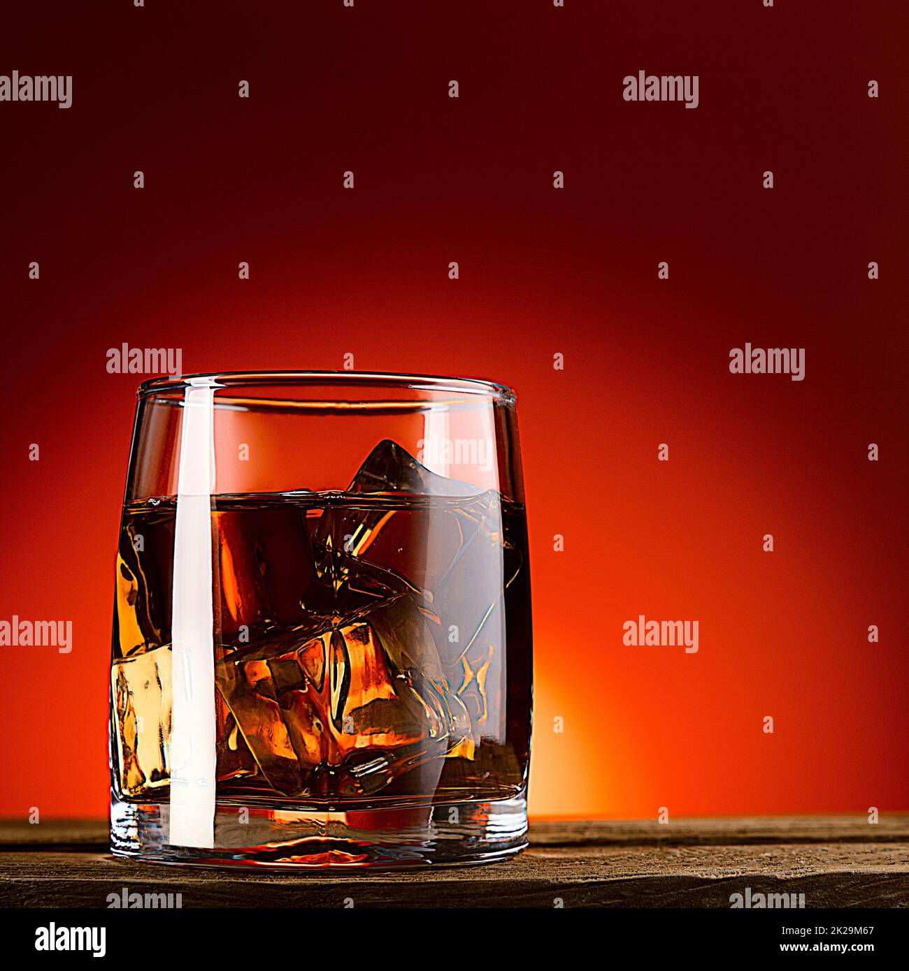 A glass of whiskey or cognac and ice cubes, close-up on a wooden table. Red background with gradient. Stock Photo