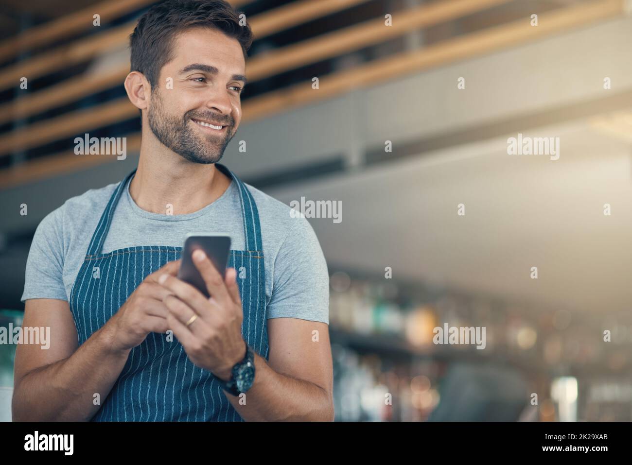 Smart baristas use the smartest tech. Shot of a young man using a mobile phone while working at a coffee shop. Stock Photo