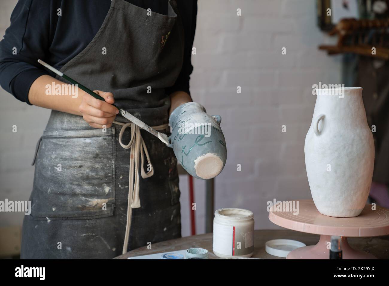 Female artisan in apron creates ceramic tableware painting white vase with brush in workshop Stock Photo