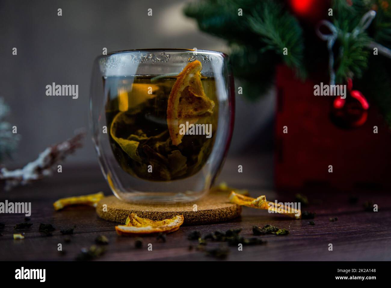 A transparent cup of tea stands on a dark table, on a wooden stand, dried orange slices float in the cup, citrus slices lie next to it, tea is scattered, a photo in a dark key Stock Photo