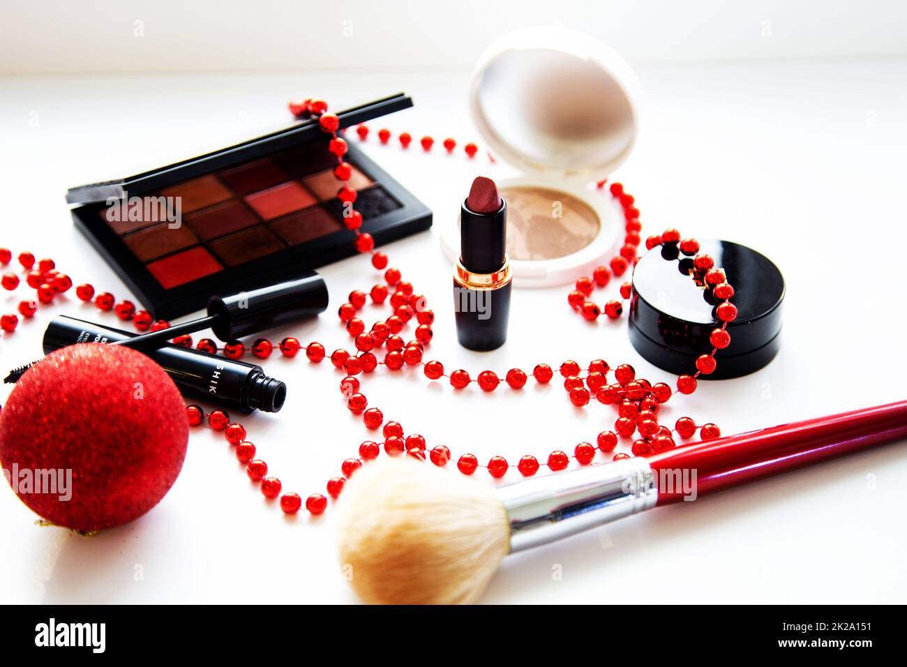 Powder and bronzer, eye shadow, lipstick, blush and brush of different brand brands lie on a white background surrounded by red Christmas balls. Stock Photo