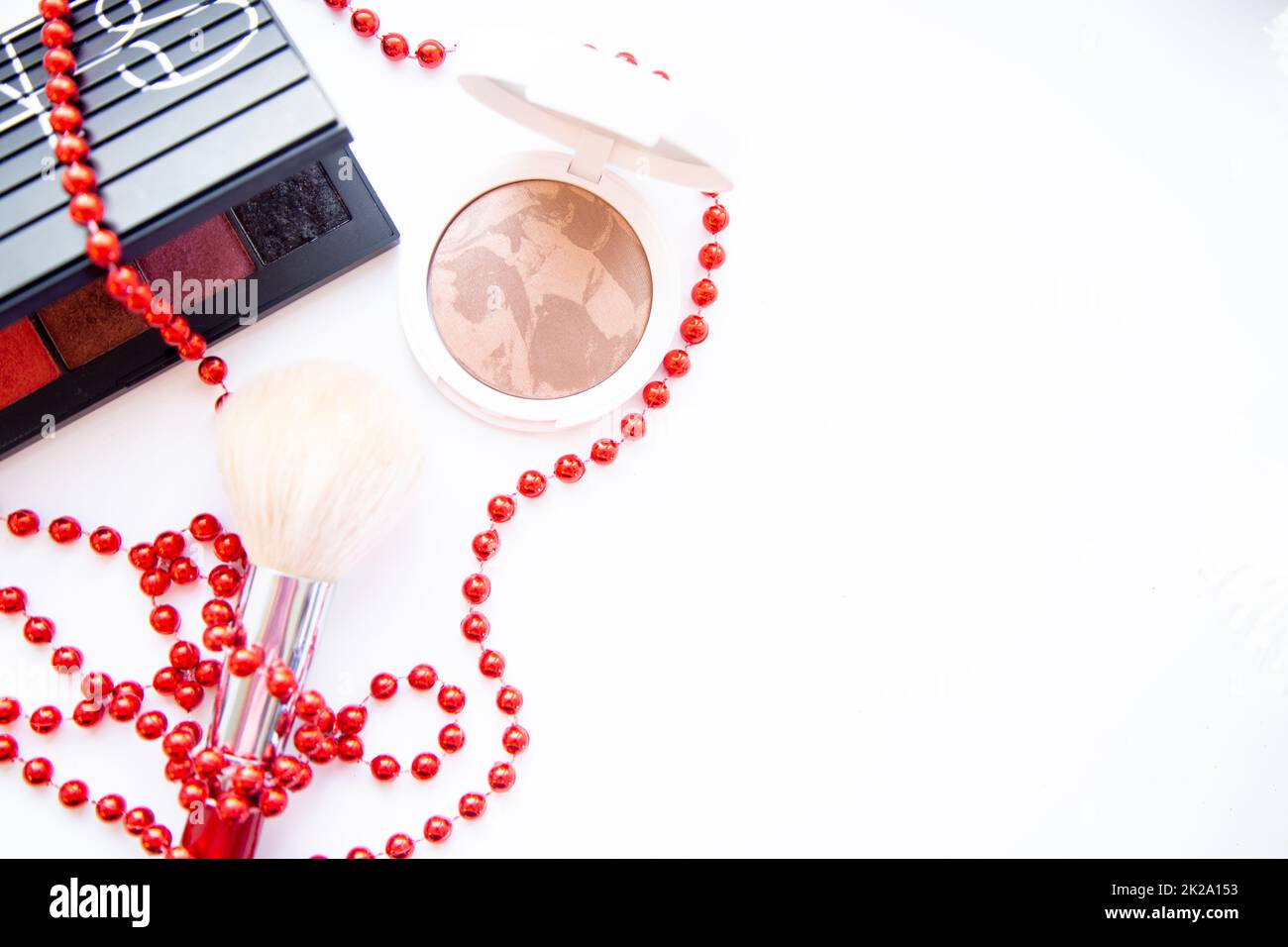 A white pillbox with powder and bronzer, a black package with eye shadow and a brush lie on a white background, they are entwined with red beads, with a place for text Stock Photo