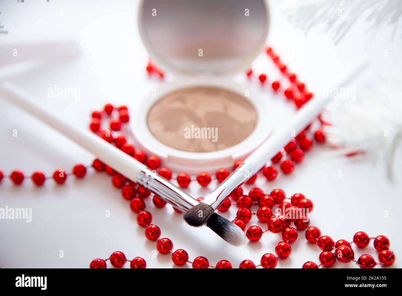 A white pillbox with powder and bronzer lies on a white background, thin makeup brushes and bright red beads lie in front. Stock Photo