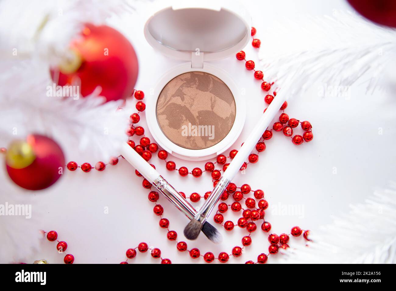 A white pillow with powder and bronzer lies on a white background among a white fluffy Christmas tree with red toys, thin makeup brushes and bright red beads lie in front. Stock Photo