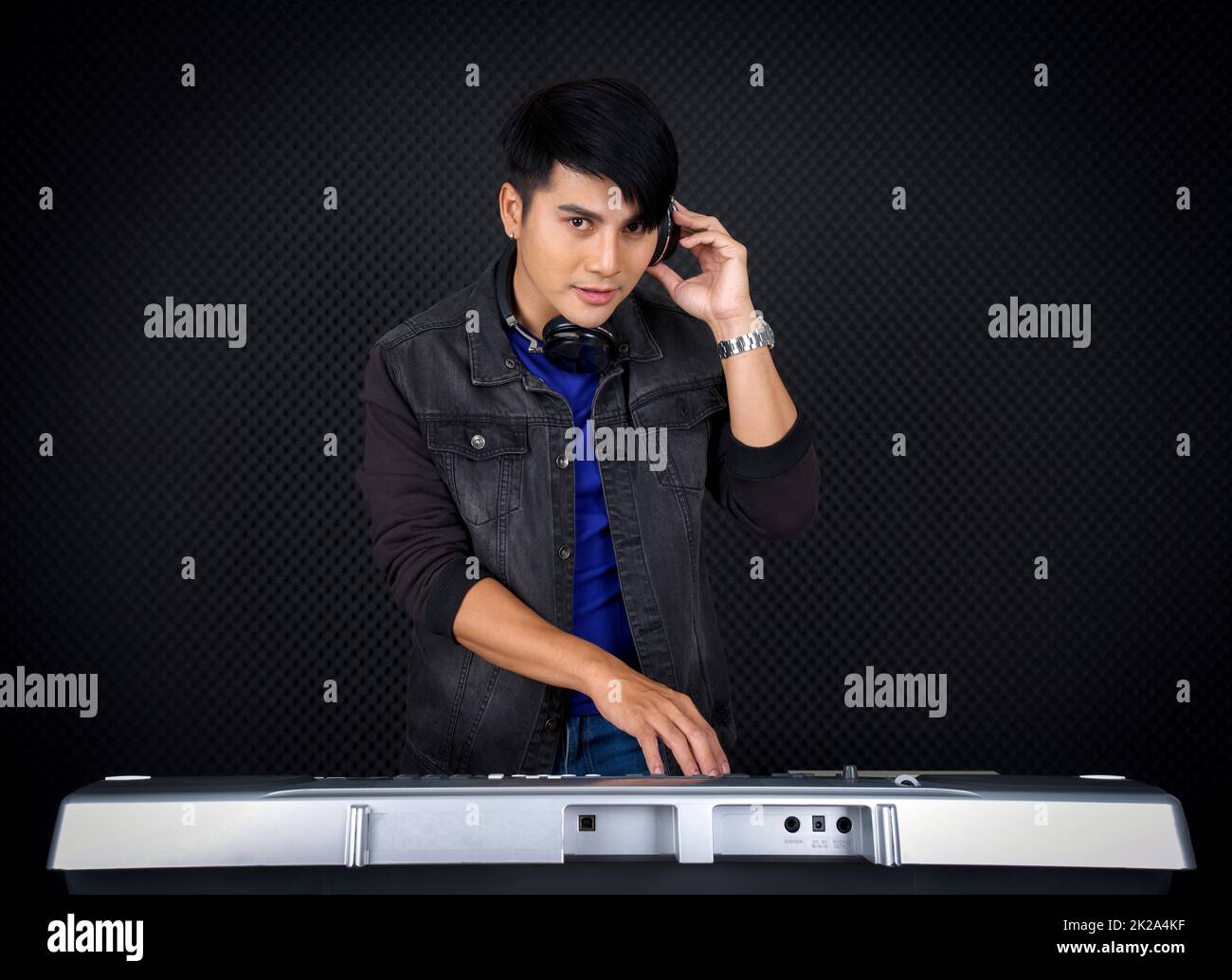 Young asian man with headphones playing an electric keyboard in front of black soundproofing wall. Musician producing music in professional recording studio. Stock Photo