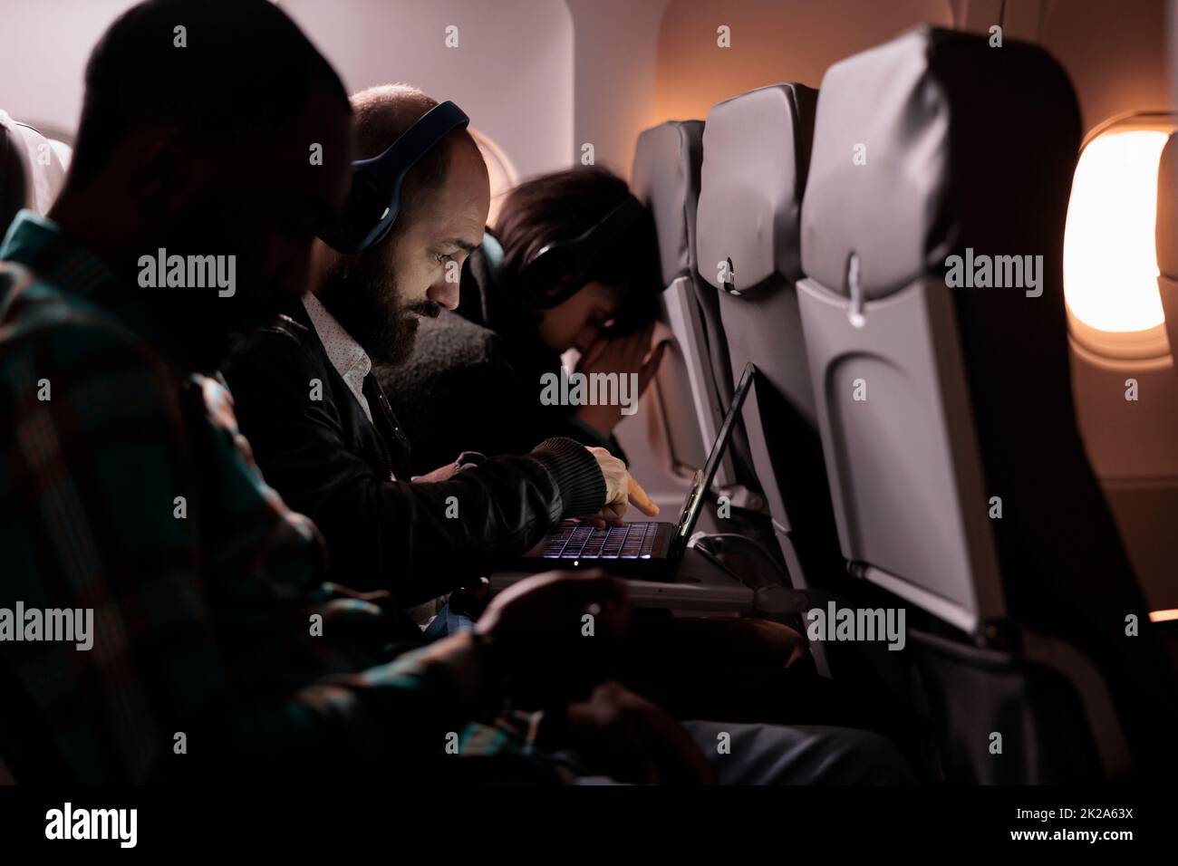 Multiethnic group of people flying in economy class together to arrive at destination, man using laptop computer during international flight. Tourists travelling abroad by airplane. Stock Photo