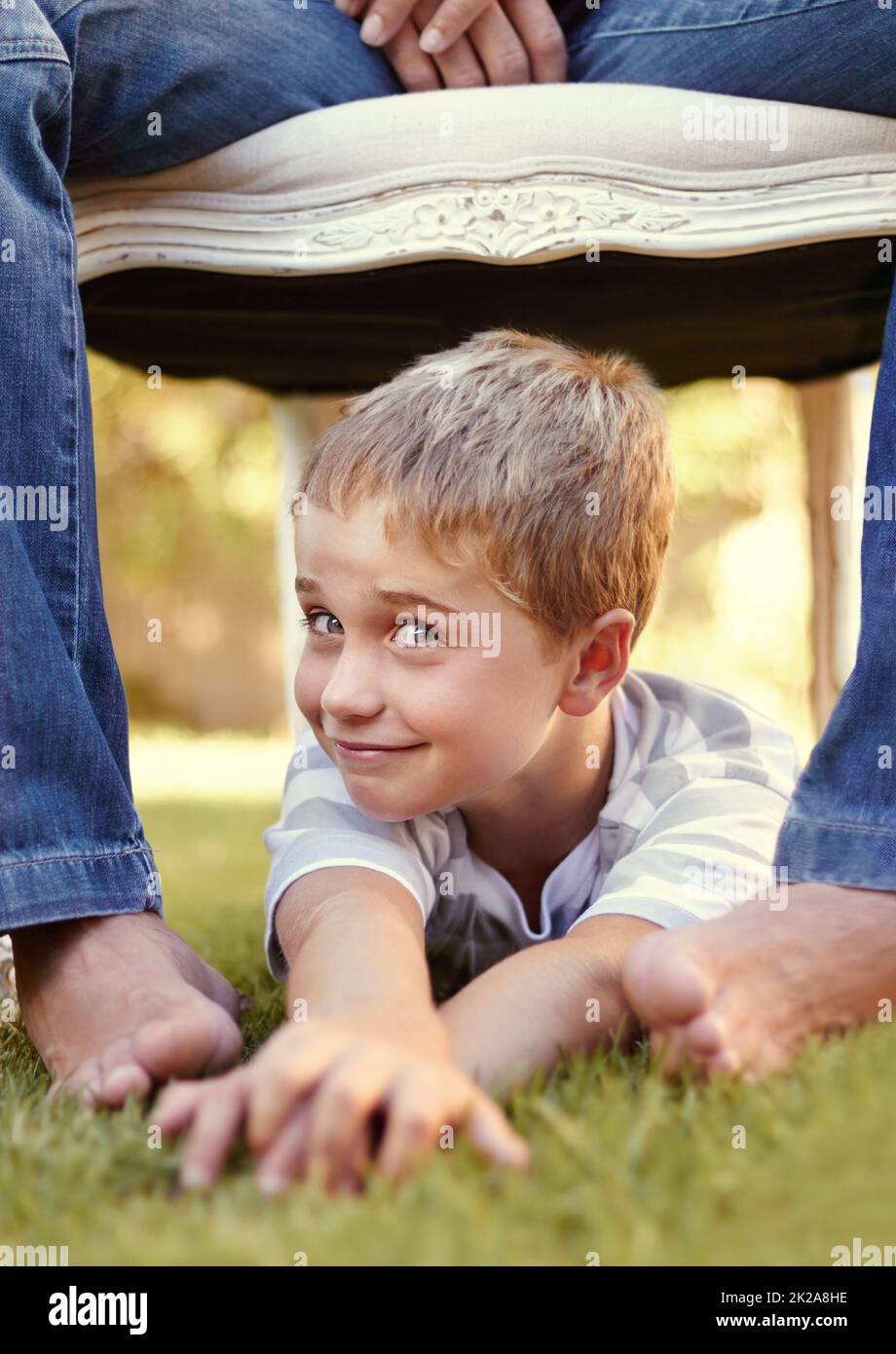 Think I should tickle his feet. Shot of a cute little boy lying on the grass underneath a chair which his father is sitting on. Stock Photo
