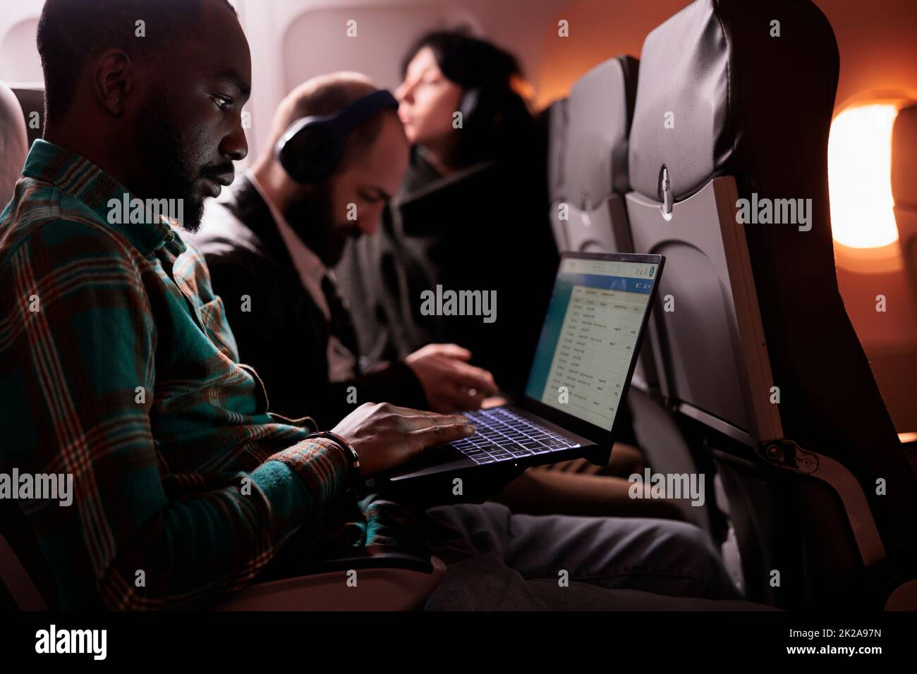 Diverse group of passengers flying in commercial class together to arrive at destination, man using laptop computer during international flight. Tourists travelling abroad by airplane. Stock Photo