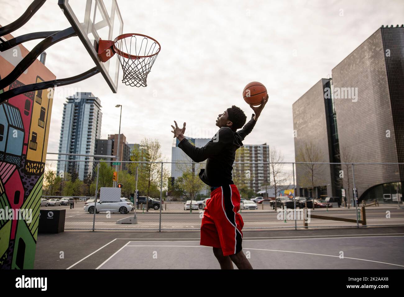 Young man shooting hoops on urban basketball court with mural Stock