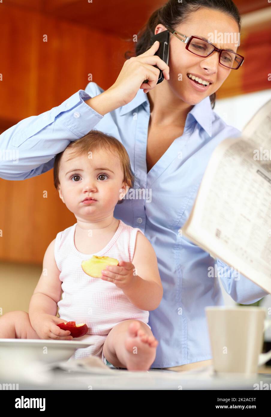 Im feeling a bit left out. Shot of a frustrated-looking single mom feeding her baby while trying to talk on the phone and holding a newspaper. Stock Photo