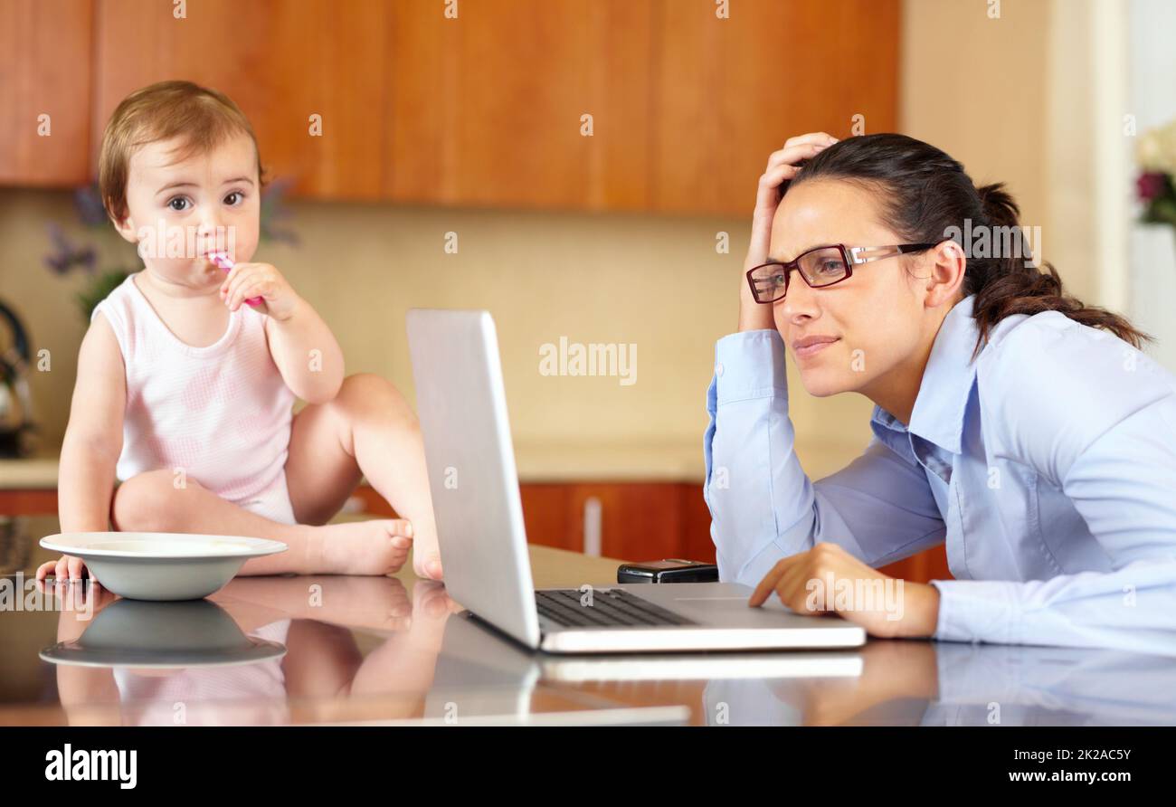 The stress of the single mom. Shot of an overworked mom working at her laptop with her baby sitting on the counter. Stock Photo