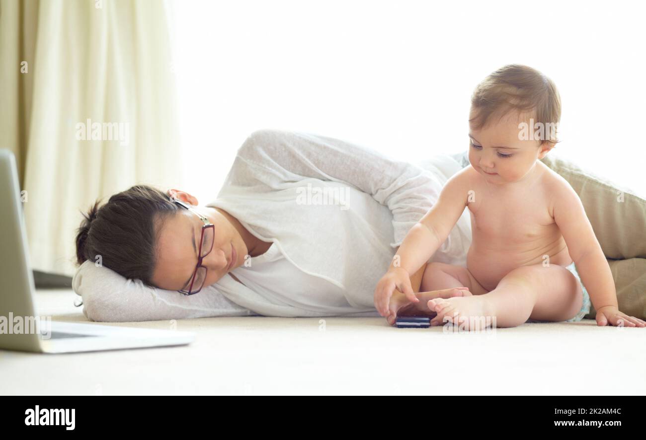 Motherhood can be exhausting. An exhausted young mother lying asleep on the floor with her baby sitting next to her. Stock Photo
