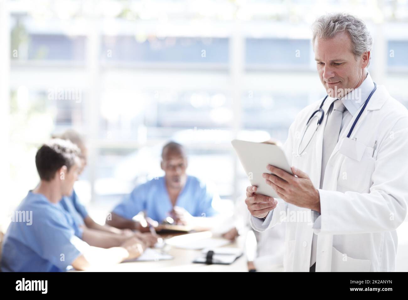 Doing his daily check. A senior doctor holding a touchpad with colleagues sitting in the background. Stock Photo
