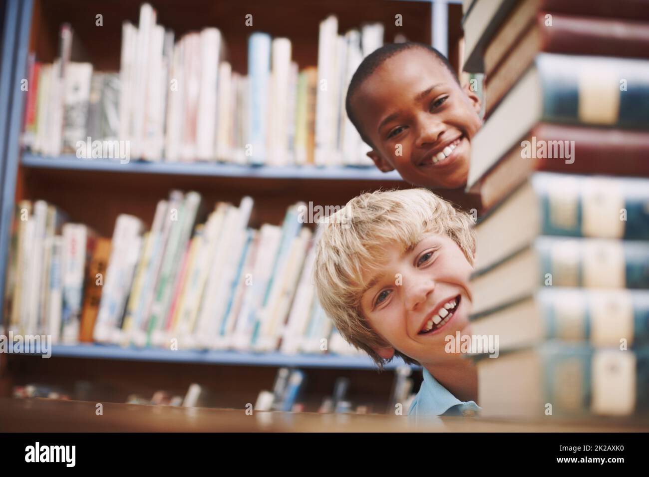 Hello. Two school friends peering around a stack of books in the library. Stock Photo