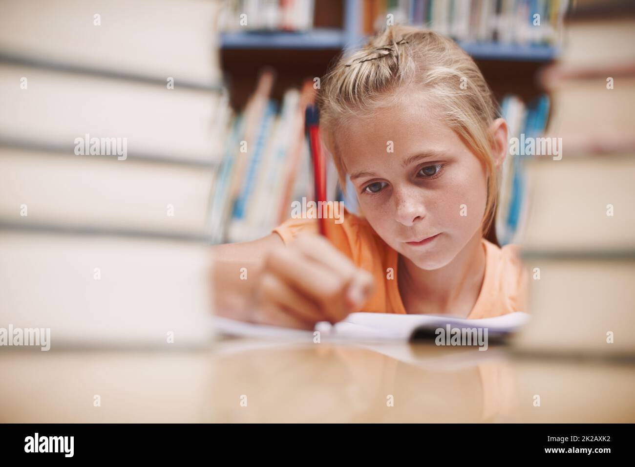 Hard at work. A cute young girl doing schoolwork while surrounded by books at the library. Stock Photo