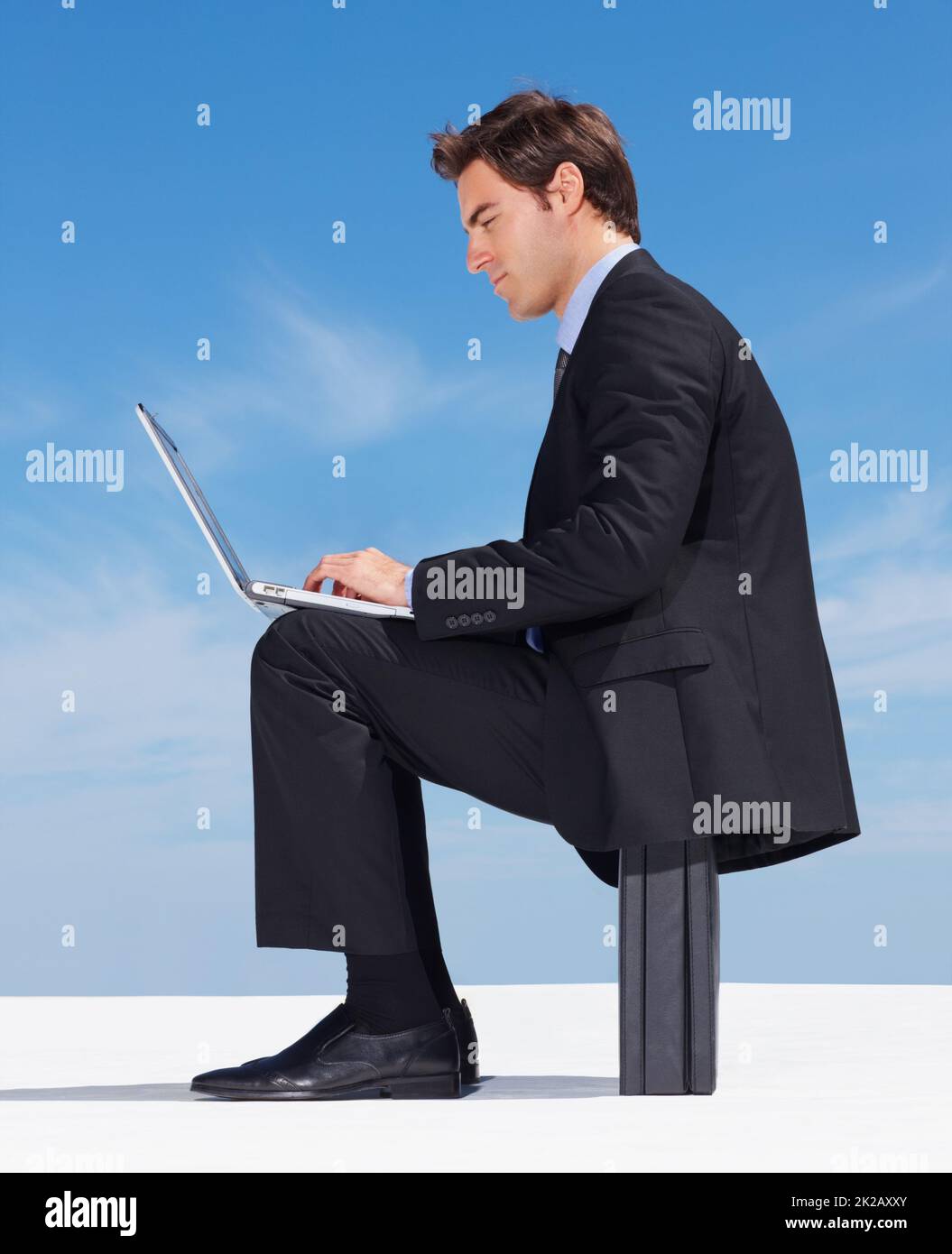 Young executive working on a laptop against clear sky. Side view of a successful young business man working on a laptop against clear sky. Stock Photo