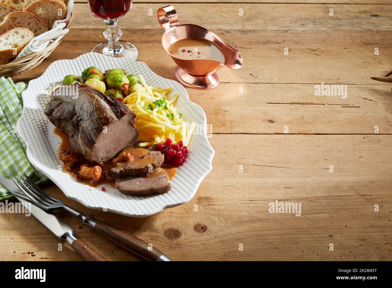 From above yummy deer venison and vegetables served on plate near sauce boat and utensils on lumber table during lunch Stock Photo