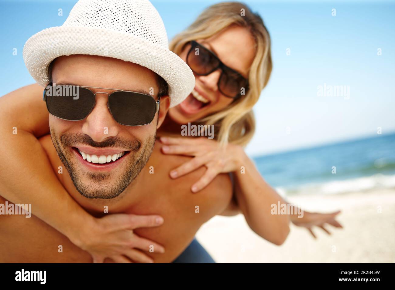 Best vacation EVER. Shot of a smiling young man giving his laughing girlfriend a piggyback on a sunny beach. Stock Photo
