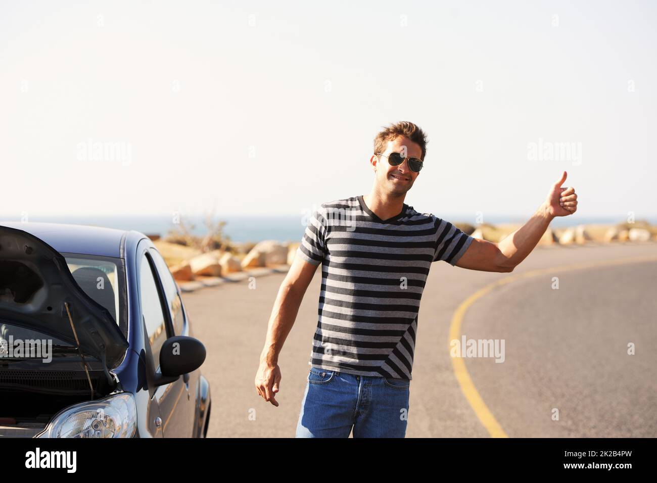 Hes hitching a ride. A handsome young man standing on the side of the road besides his broken-down car and hitching a ride. Stock Photo