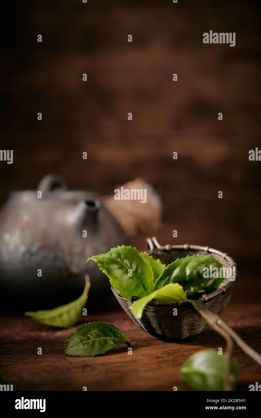 Green tea leaves for brewing in metal ladle placed on table with old teapot on blurred background in light room Stock Photo