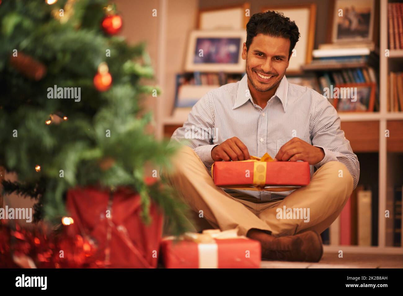 Everyone feels like a kid at christmas time. Portrait of a young man sitting next to the tree opening christmas presents. Stock Photo