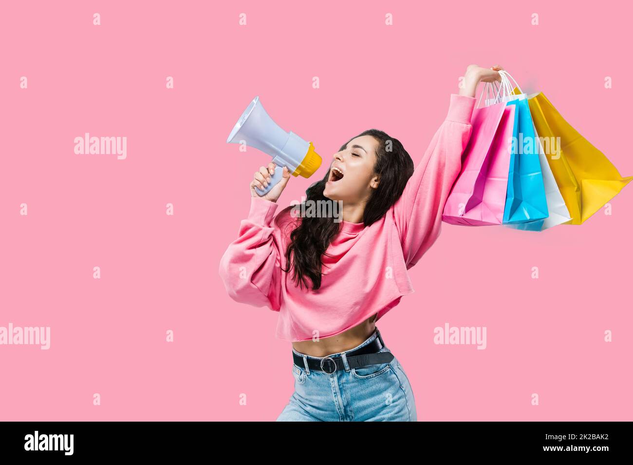 Big sales concept. Black friday. Happy trendy pretty caucasian young woman, in casual clothes, holding shopping bags, says announcement in loudspeaker, posing on isolated pink background Stock Photo