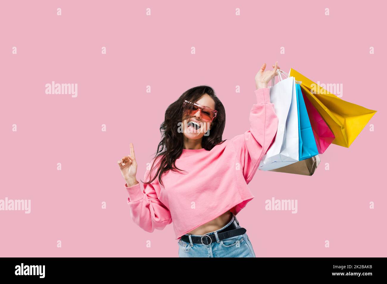 Shopping and sales concept. Cheerful trendy pretty caucasian young woman, in casual clothes and pink sunglasses, holding shopping bags, posing on isolated pink background, looks at camera, smiling Stock Photo