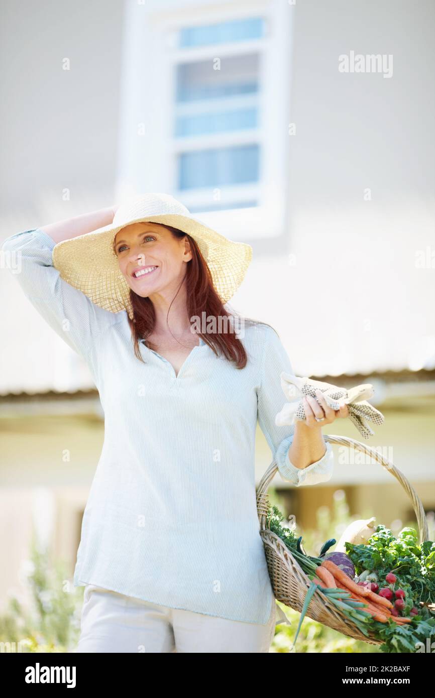 Gardening is how I relax. A beautiful woman holds her gardening gloves and a basket of freshly picked vegetables while standing in her garden. Stock Photo