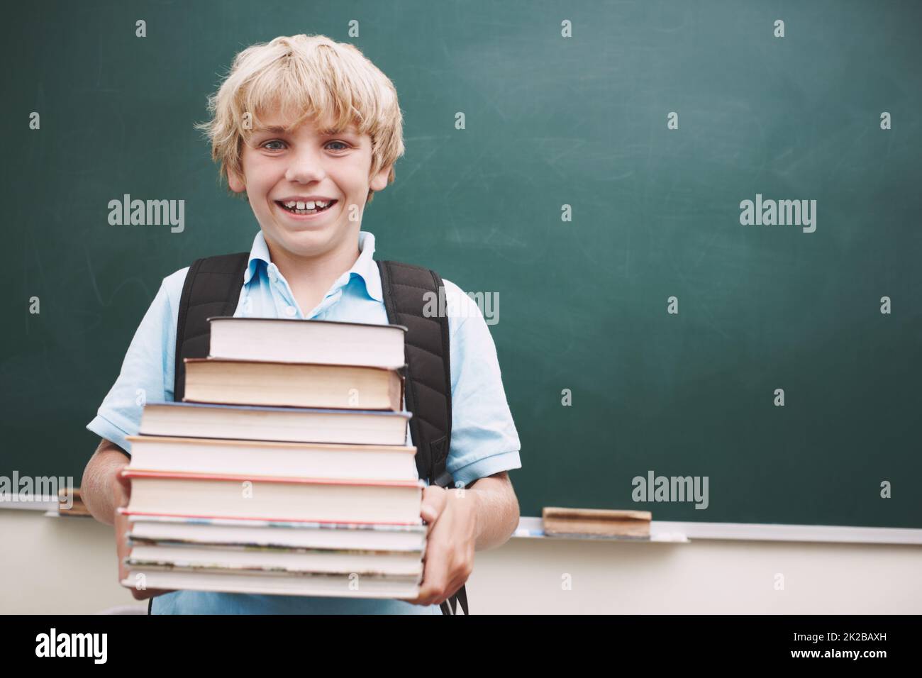 I want to pack my brain as full as possible. A cute young boy carrying a stack of books against a blackboard in the classroom. Stock Photo