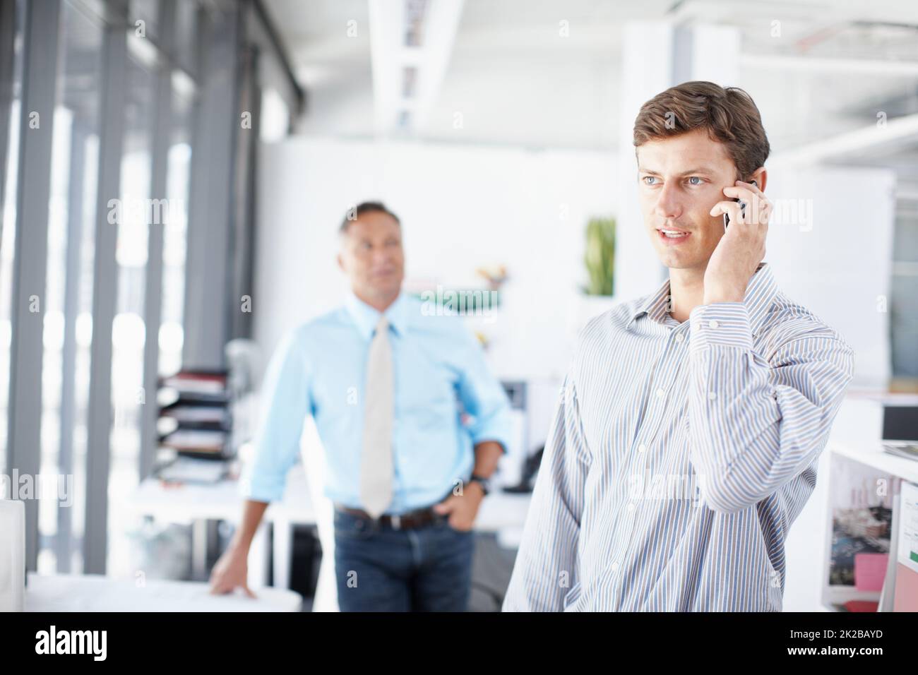 Getting updated on the design brief. A young professional talking on his cellphone as a colleague stands in the background. Stock Photo