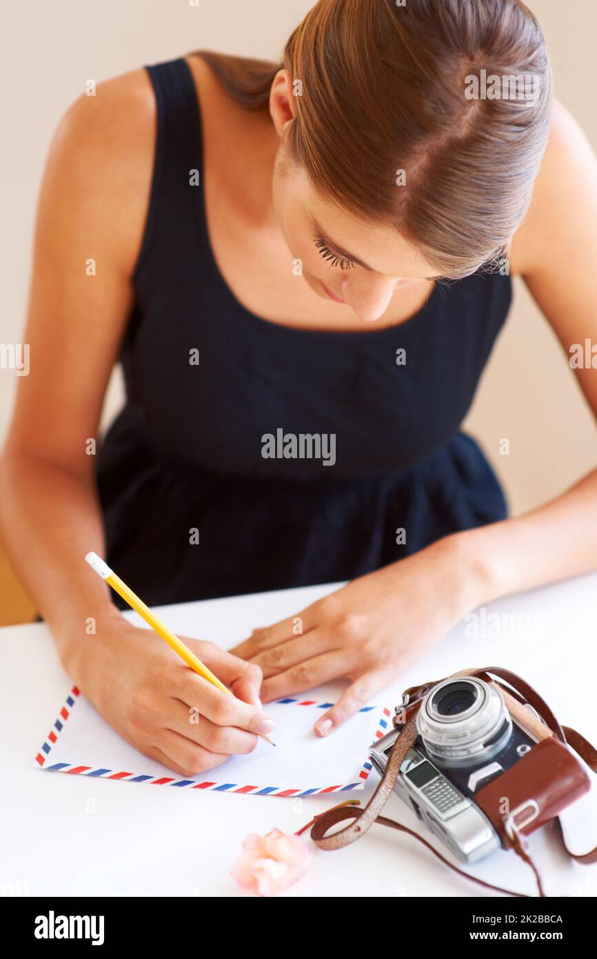 Writing home. A young woman writing an address on an envelope with a camera lying on her desk. Stock Photo