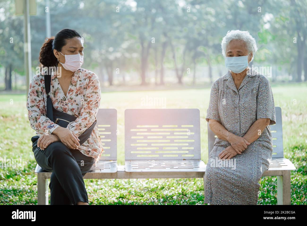 Asian elderly woman and daughter in social distancing sitting bench and wearing face mask for protect safety infection Covid-19 Coronavirus in park. Stock Photo