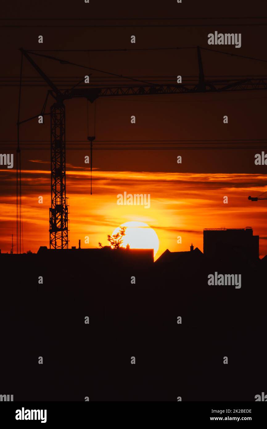 Tall construction crane silhouette in orange sky sunset shows construction site with engineering for modern buildings and city development as architectural teamwork for skyscrapers high voltage lines Stock Photo