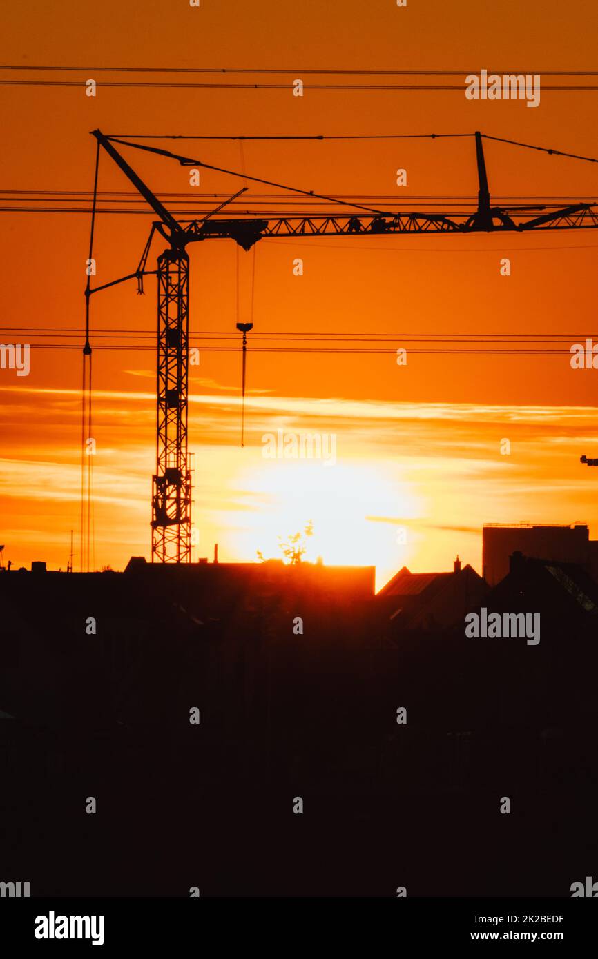 Tall construction crane silhouette in orange sky sunset shows construction site with engineering for modern buildings and city development as architectural teamwork for skyscrapers high voltage lines Stock Photo