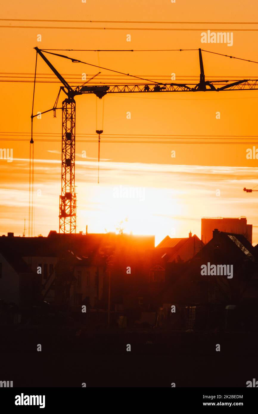 Tall construction crane silhouette in orange sky sunset shows construction site with engineering for modern buildings and city development as architectural teamwork for skyscrapers high voltage lines Stock Photo